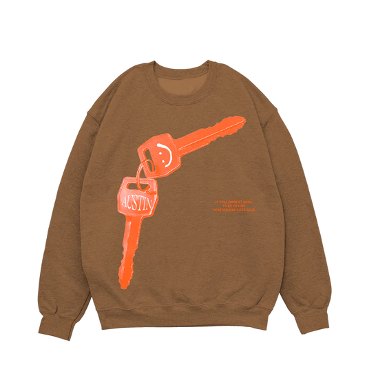 "Tan crewneck sweatshirt featuring a large orange key graphic with the word 'Austin' and a smiley face keychain, along with text 'If y'all weren't here, I'd be crying. Post Malone © 2023 Tour' in orange."